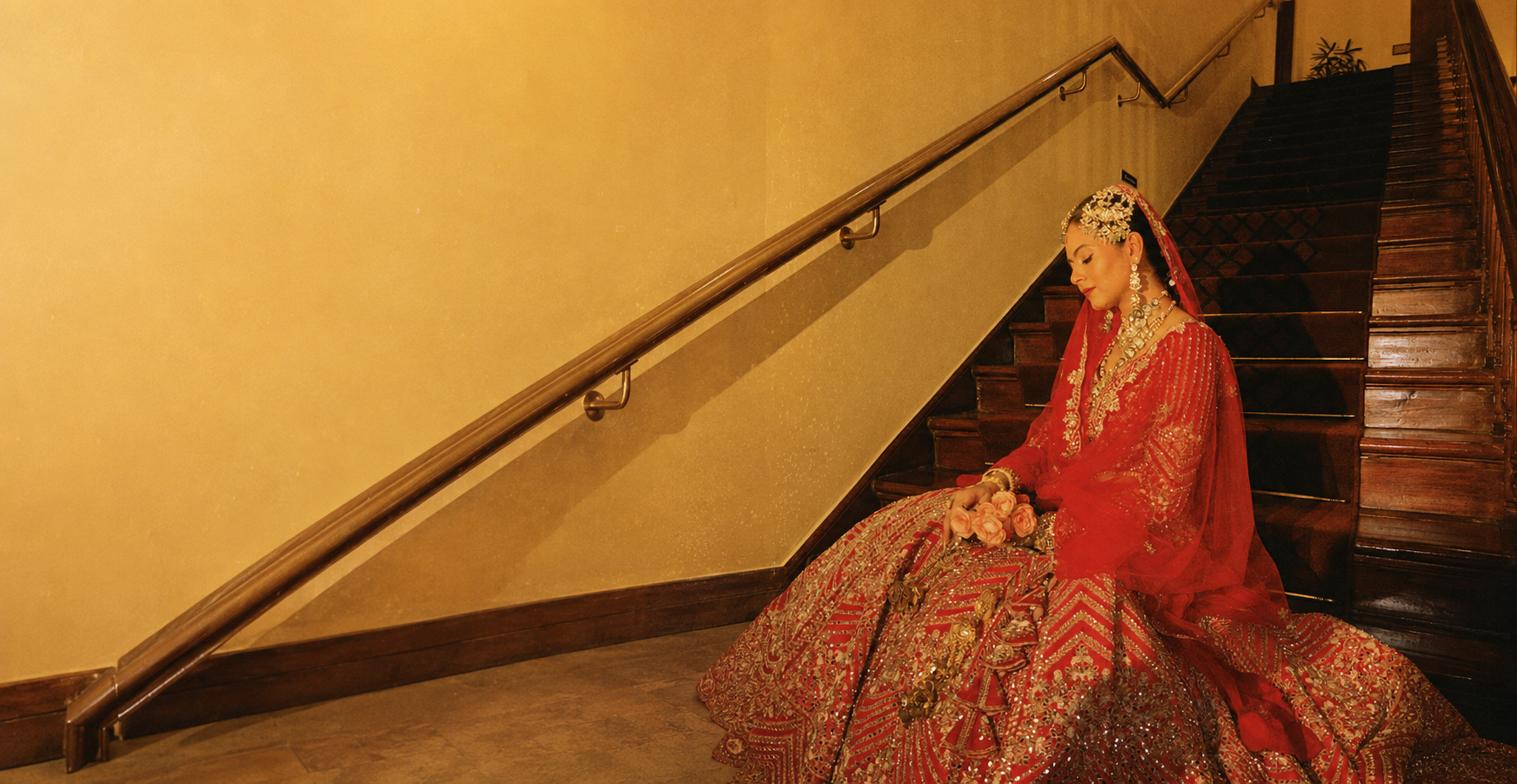 Bride in a red lehenga seated on a staircase, makeup by The Makeup Whisperers